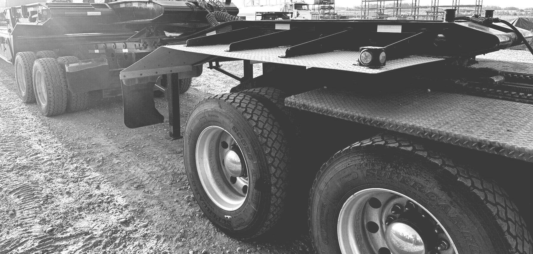 Lowbed trailer decks and heavy tires on a gravel yard, with trucks and equipment standing behind them