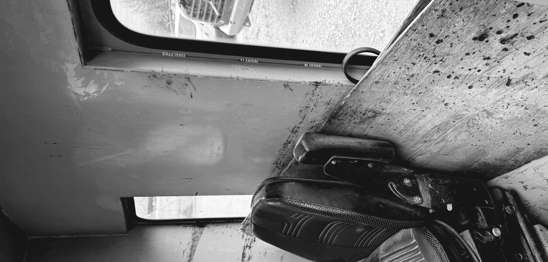 Inside a working cab: the operator's seat, a worn steel deck and an open door looking out across the yard to another machine